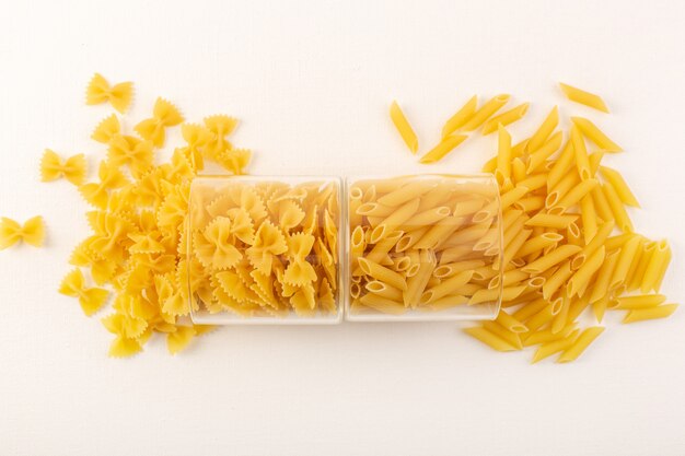 A top view raw pasta dry italian yellow pasta inside transparent plastic bowls and spread on the white background italian food meal