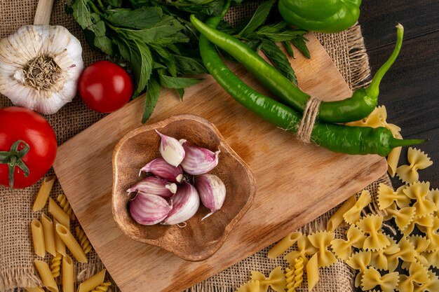 Top view of raw pasta with garlic and chili peppers on a cutting board with tomatoes and a bunch of mint on a beige napkin