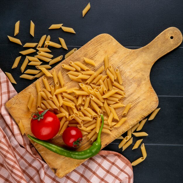 Top view of raw pasta with tomatoes and chilli pepper on a cutting board on a black surface