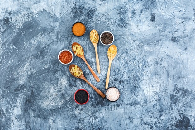 Top view raw pasta in wooden spoons with spices on grey plaster background. horizontal