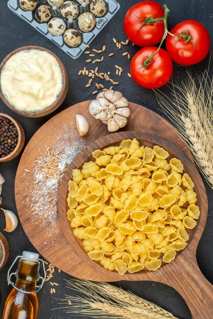 Top view of raw pastas in a brown bowl garlics rice on wooden board flour eggs spikes fallen oil bottle peppers tomatoes with stemon black background