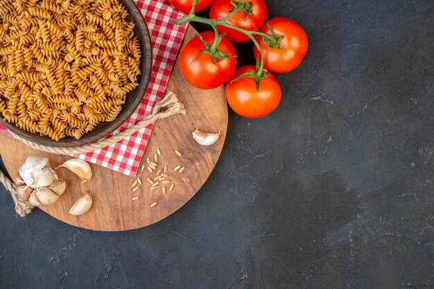 Top view of raw pastas in a brown bowl on red stripped towel garlics rice on round wooden board tomatoes rope on the right side on black table