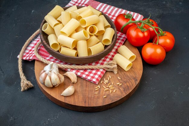 Top view of raw pastas inside and outside a brown bowl on red stripped towel garlics rice on round wooden board tomatoes rope on black background