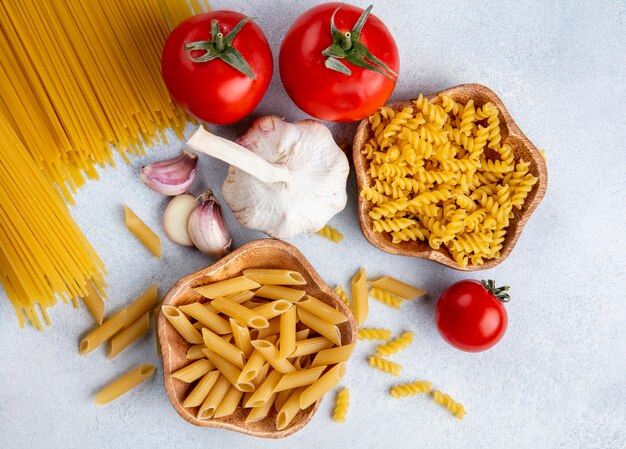 Top view of raw spaghetti with raw pasta in bowls with garlic and tomatoes on a gray surface