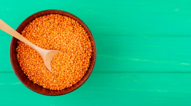 Top view of raw uncooked red lentils in a bowl with wooden spoon on green background with copy space