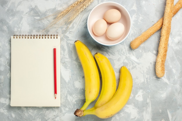 Top view raw whole eggs inside little plate with bun bread and bananas on light-white desk raw food meal breakfast fruit bread