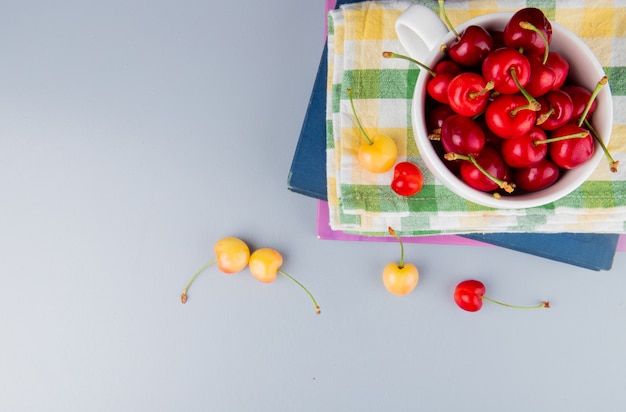 Top view of red cherries in cup on cloth and books with yellow cherries on blue surface with copy space