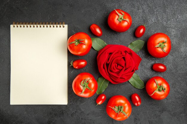 Top view red and cherry tomatoes around a towel in rose shape and bay leaves and a notebook on dark ground