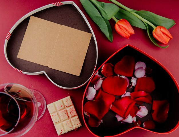 Top view of red color tulip flowers with heart shaped gift box with an open postcard and a box filled with rose petals and white chocolate with a glass of wine on red background