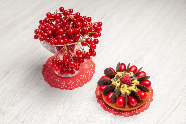 Top view red currant in a crystal glass and berry cake on the red oval lace doily on the white wooden table