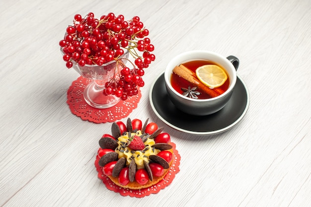 Top view red currant in a crystal glass on the red oval lace doily a cup of lemon cinnamon tea and berry cake on the white wooden table