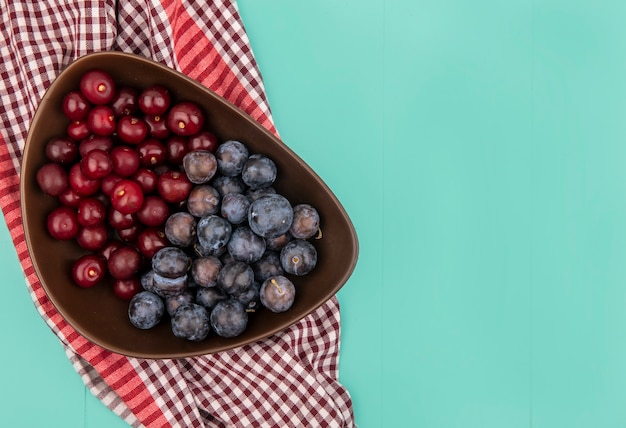 Top view of red delicious cherries with dark purple sloes on a wooden bowl on a checked tablecloth on a blue background with copy space