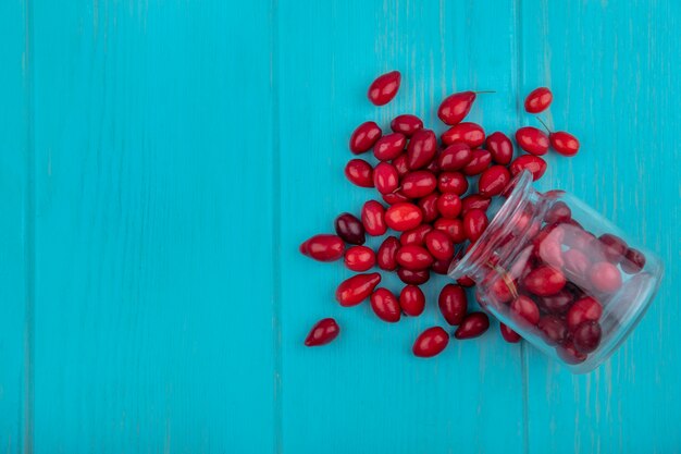 Вид сверху красных и свежих ягод кизила, падающих из стеклянной банки на синем деревянном фоне с копией пространства
