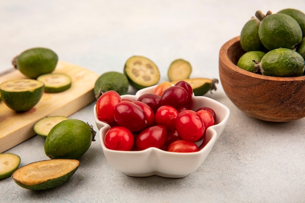 Top view of red fresh cornelian cherries on a bowl with feijoas on a wooden bowl on a grey wall