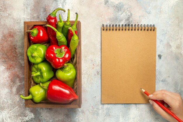 Free photo top view red and green peppers hot peppers in wooden box notebook red pencil in female hand on nude surface