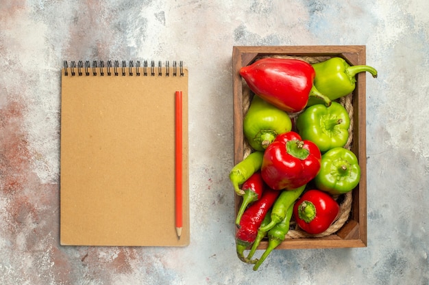 Free photo top view red and green peppers hot peppers in wooden box a notebook red pencil on nude surface