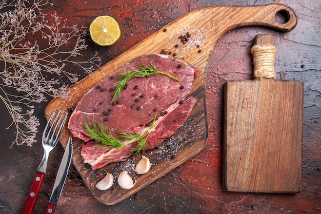 Top view of red meat on wooden cutting board garlic fork and knife on dark background