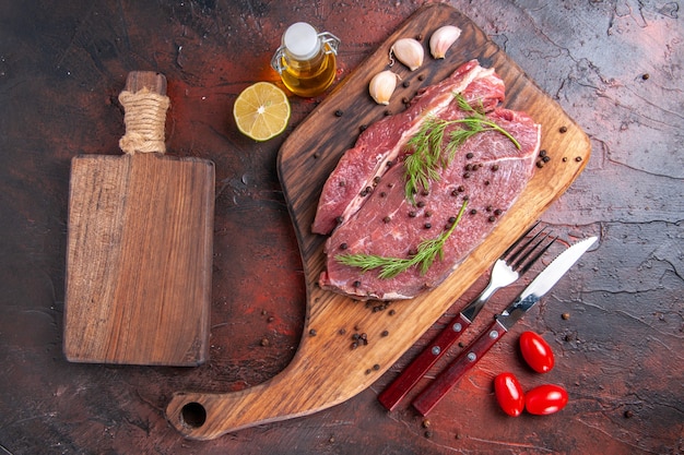 Top view of red meat on wooden cutting board and garlic green pepper oi bottle fork and knife on dark background