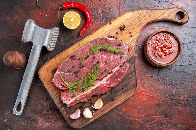 Вид сверху красного мяса на деревянной разделочной доске и чеснока, вилки и ножа для бутылки с маслом зеленого перца на темном фоне кадра