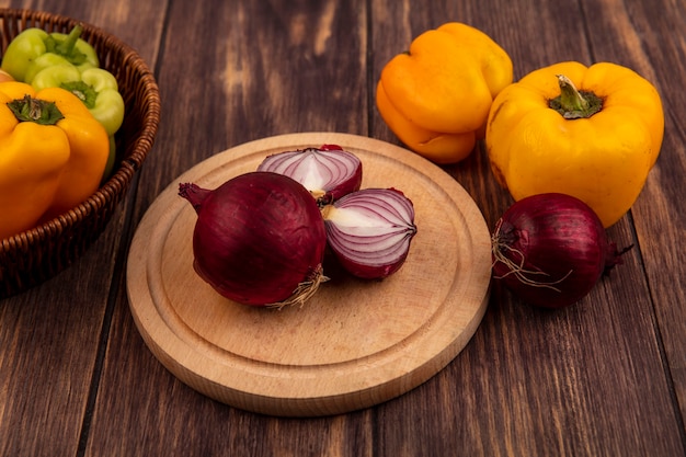 Top view of red onions on a wooden kitchen board with bell peppers on a bucket with yellow peppers isolated on a wooden wall