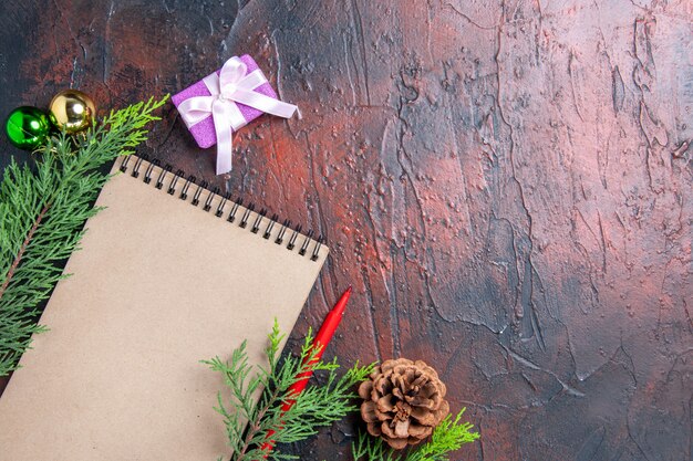 상위 뷰 빨간 펜 노트북 소나무 나뭇 가지 크리스마스 트리 볼 장난감 및 여유 공간이있는 어두운 빨간색 표면에 선물