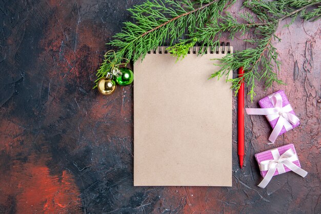 Top view red pen a notepad pine tree branches xmas tree toys and gifts on dark red surface free space