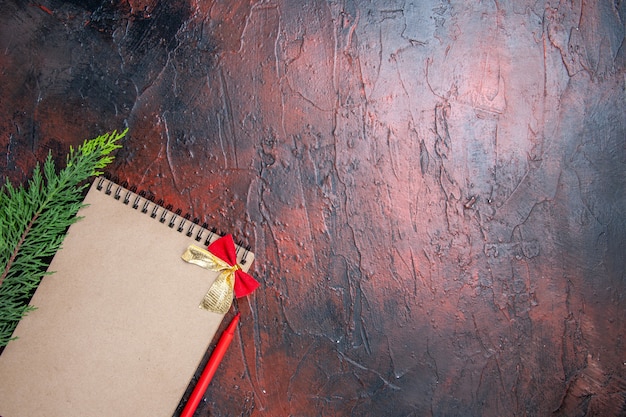 Top view red pen a notepad with little bow a pine tree branch at the bottom left of dark red surface with copy space