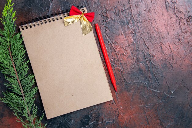 Top view red pen a notepad with little bow a pine tree branch on dark red surface copy space