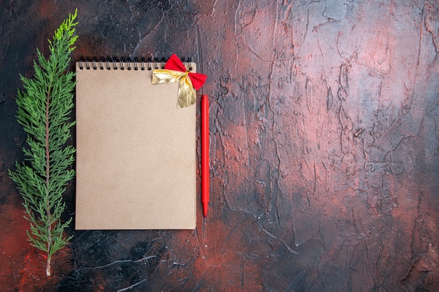 Top view red pen a notepad with little bow a pine tree branch on dark red surface with copy place