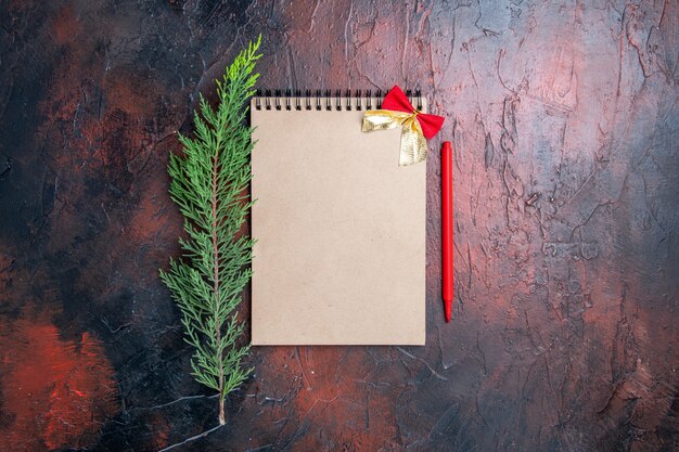 Top view red pen a notepad with little bow a pine tree branch on dark red surface with copy space