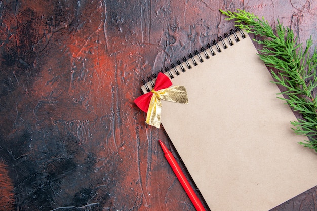 Top view red pen a notepad with little bow a pine tree branch at the right of dark red surface with copy space