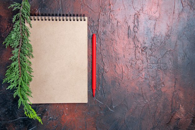 Top view red pen a notepad with small bow a pine tree branch on dark red surface copy space