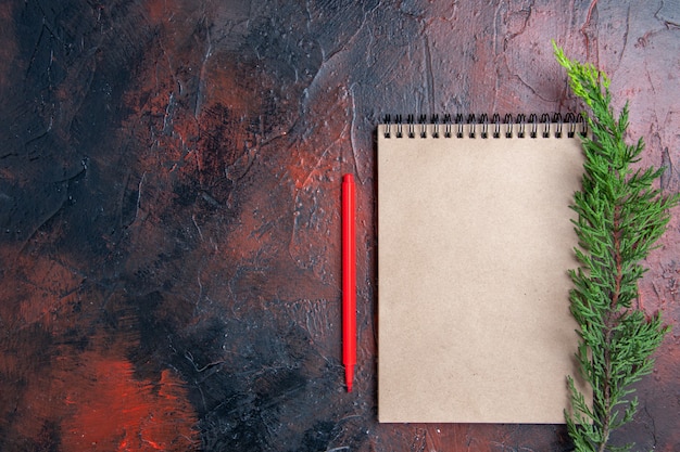 Top view red pen a notepad with small bow a pine tree branch on dark red surface with copy place