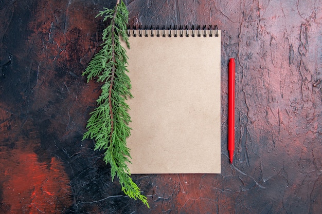 Top view red pen a notepad with small bow a pine tree branch on dark red surface with copy space