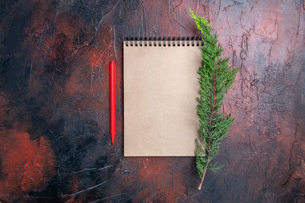 Top view red pen a notepad with small bow a pine tree branch on dark red surface with copy space