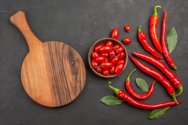 Top view red peppers and pay leaves and a bowl of cherry tomatoes and an oval chopping board on black table with free space