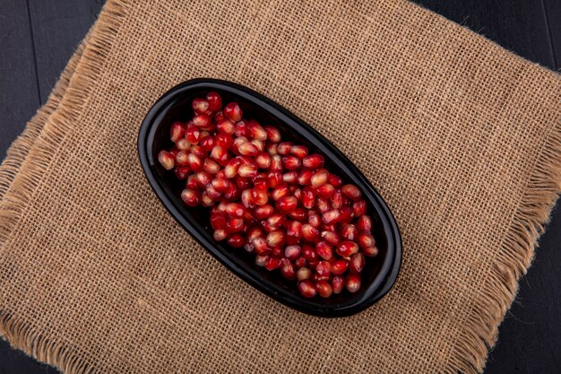 Free photo top view of red pomegranate seeds in a black plate on sack cloth surface