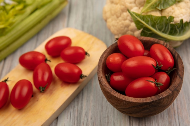 Top view of red skinned tomatoes on a wooden kitchen board with tomatoes on a wooden bowl with cauliflower and celery isolated on a grey wooden surface