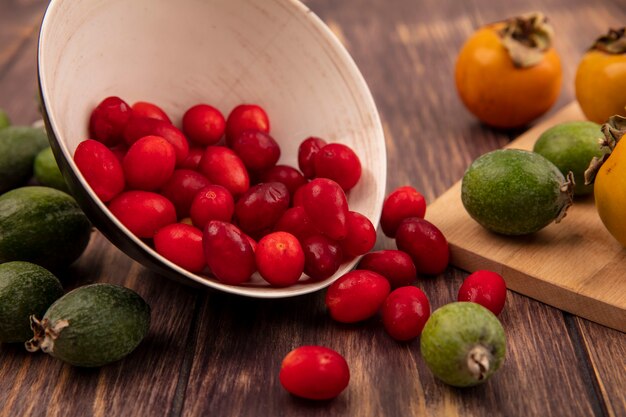 Free photo top view of red sour cornelian cherries falling out of a bowl with persimmon and feijoas isolated on a wooden wall