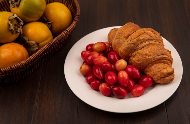 나무 벽에 양동이에 감 과일과 흰 접시에 크로와 붉은 신 산수유 체리의 상위 뷰
