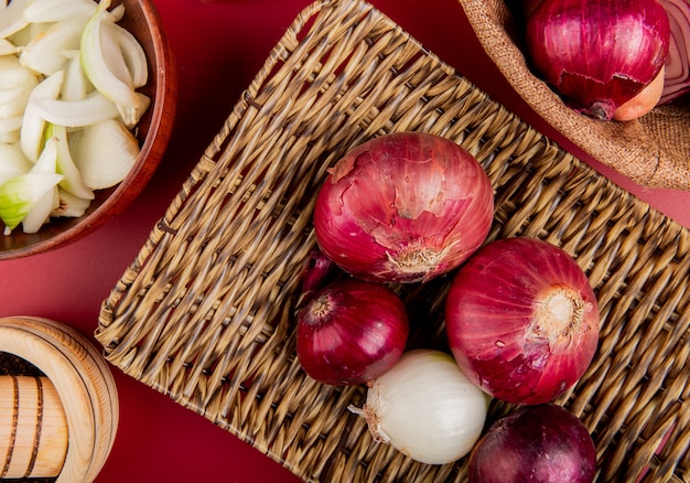 Top view of red and white onions in basket plate with sliced white one in bowl and black pepper seeds on red