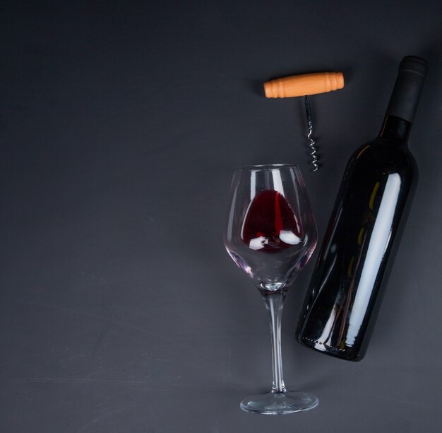 Top view of red wine in lying glass and bottle with corkscrew on black with copy space