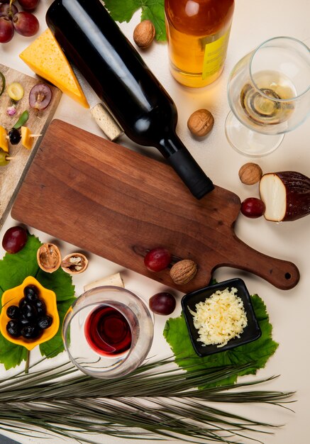 top view of red wine and white wine with olive walnut grape cutting board shredded parmesan cheese on white table decorated with leaves