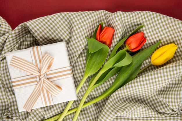 Top view of red and yellow color tulips and a present box tied with bow on plaid fabric on red table