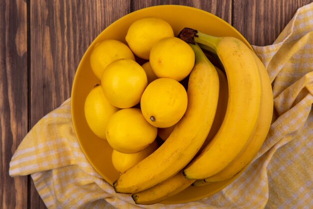 木製の壁にバナナと黄色のチェックの布の上の黄色のプレート上の豊富なビタミンレモンの上面図