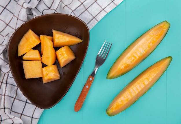 Top view of ripe delicious cantaloupe melon slices on a brown bowl with fork on checked tablecloth on blue surface