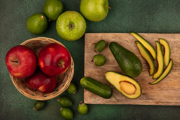 Top view of ripe fresh avocado with slices on a wooden kitchen board with red apples on a bucket with limes feijoas and green apples isolated on a green surface