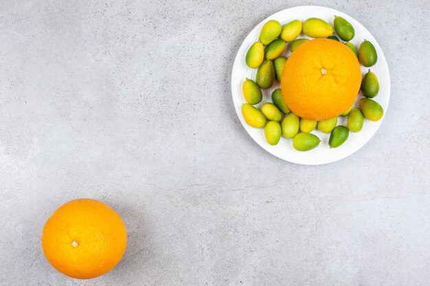 Top view of ripe orange with pile of kumquats on white plate.