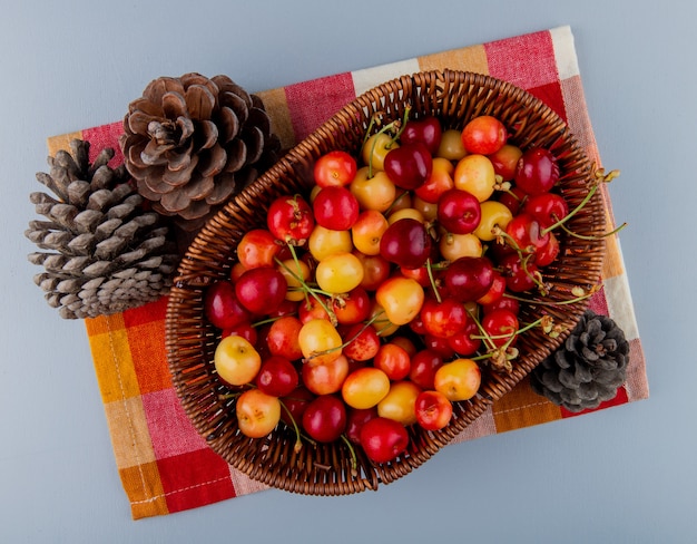 Free photo top view of ripe rainier cherriesin a wicker basket and cones on plaid napkin on grey