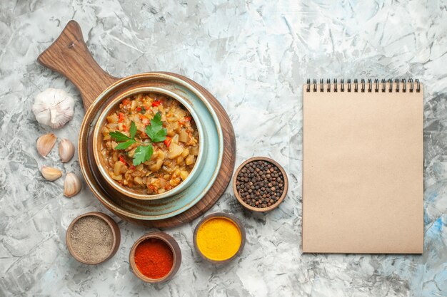 Top view of roasted eggplant salad in bowl on chopping board and different spices in bowls a notebook on grey surface